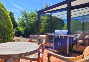 Eine Außenpatio mit Holztisch und -stühlen, einem Grill und einem gläsernen Anbau mit Blick ins grüne Umfeld.