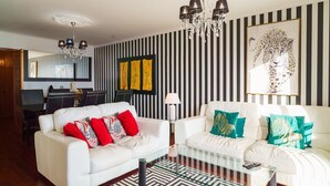 A living room with two white sofas, a glass coffee table, a striped wall, and a chandelier. - GuestReady - Breathtaking seaview in Caniço (Caniço)