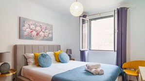 A bedroom with a bed, a painting, a window with curtains, a chair, and a lamp. - GuestReady - Breathtaking seaview in Caniço (Caniço)