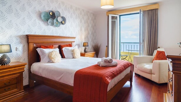 A hotel room with a large bed, wooden headboard, a balcony with a view, and a small table with a lamp. - GuestReady - Breathtaking seaview in Caniço (Caniço)
