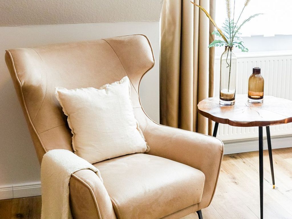 A beige leather armchair with a pillow, a small round table with two bottles and a plant, and a window with curtains.