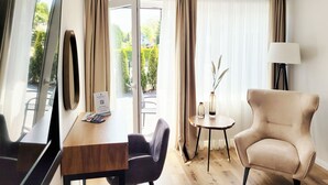 Ein Hotelzimmer mit einem Holztisch, zwei Stühlen, einem runden Beistelltisch mit einer Vase, einer Lampe und Blick auf Grün durch große Fenster.