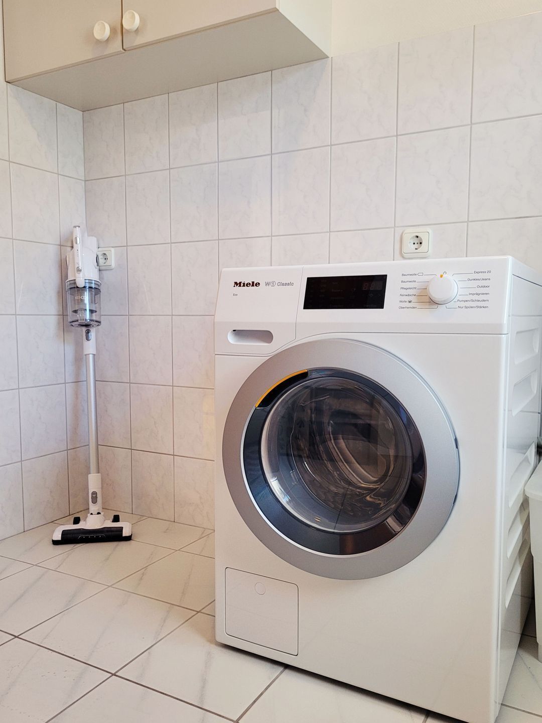 A modern, compact washing machine in a clean, tiled bathroom with a handheld vacuum cleaner.