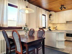 Un coin repas avec une table et des chaises en bois, un luminaire suspendu, et une cuisine dotée de meubles et d’appareils électroménagers blancs.