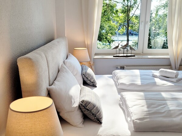 A hotel room with a bed, pillows, a lamp, a window with a view of trees, and a radiator. - A new gem on the waterfront: first-class furnishings and fantastic views (Neustadt in Holstein)