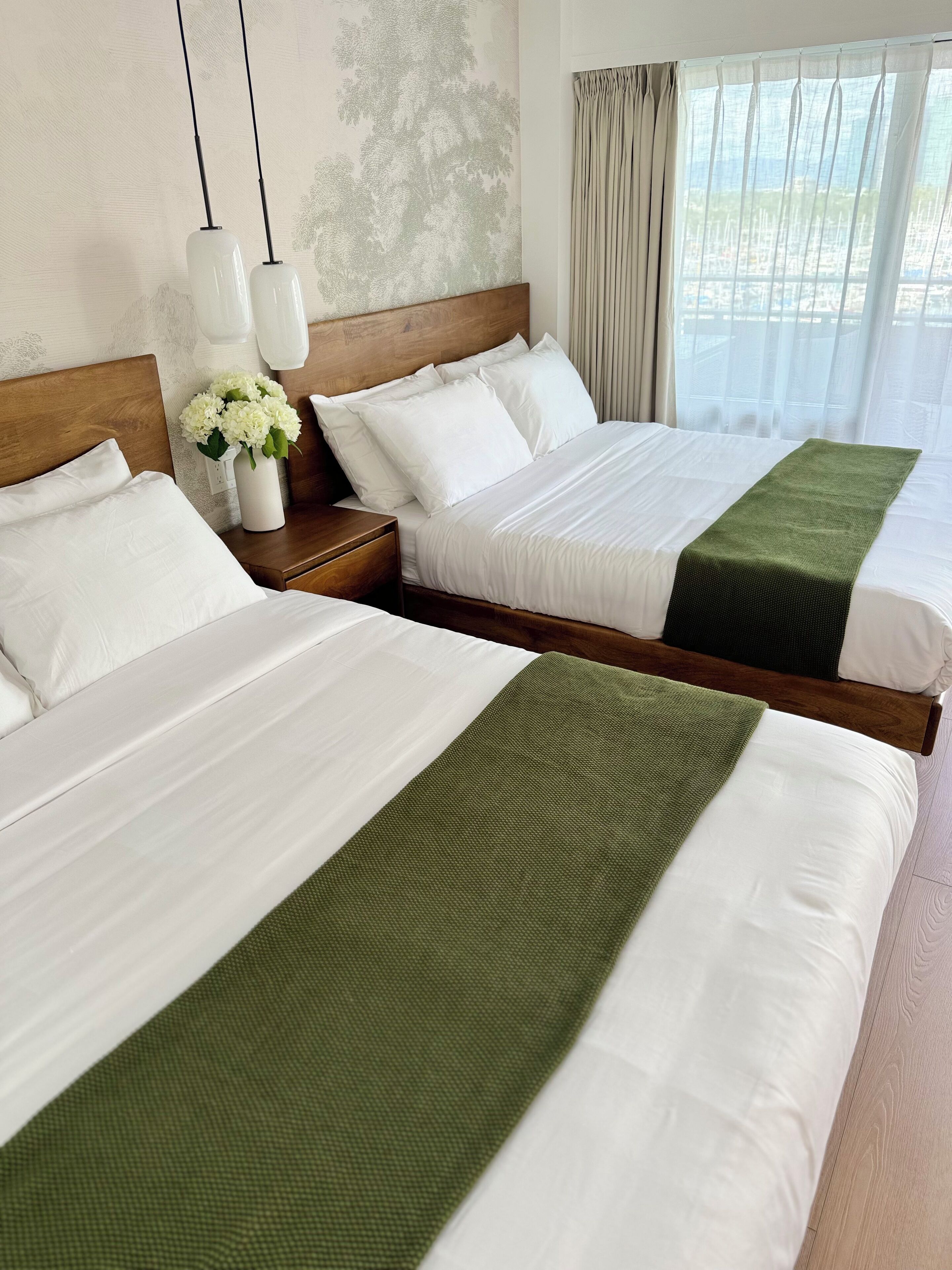 A hotel room with two beds, white bedding, a wooden headboard, a green throw blanket, a vase with white flowers, and a view of the city through sheer curtains.