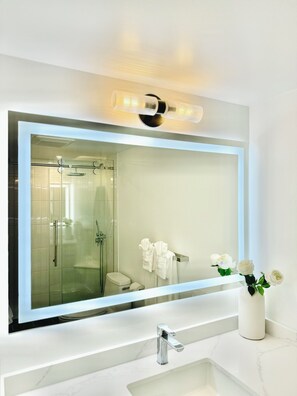 Une salle de bain moderne avec un grand miroir, une douche à parois de verre, un lavabo blanc et un vase rempli de fleurs blanches.
