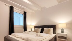 A modern hotel room with a large bed, two bedside tables with lamps, a window with curtains, and a radiator.