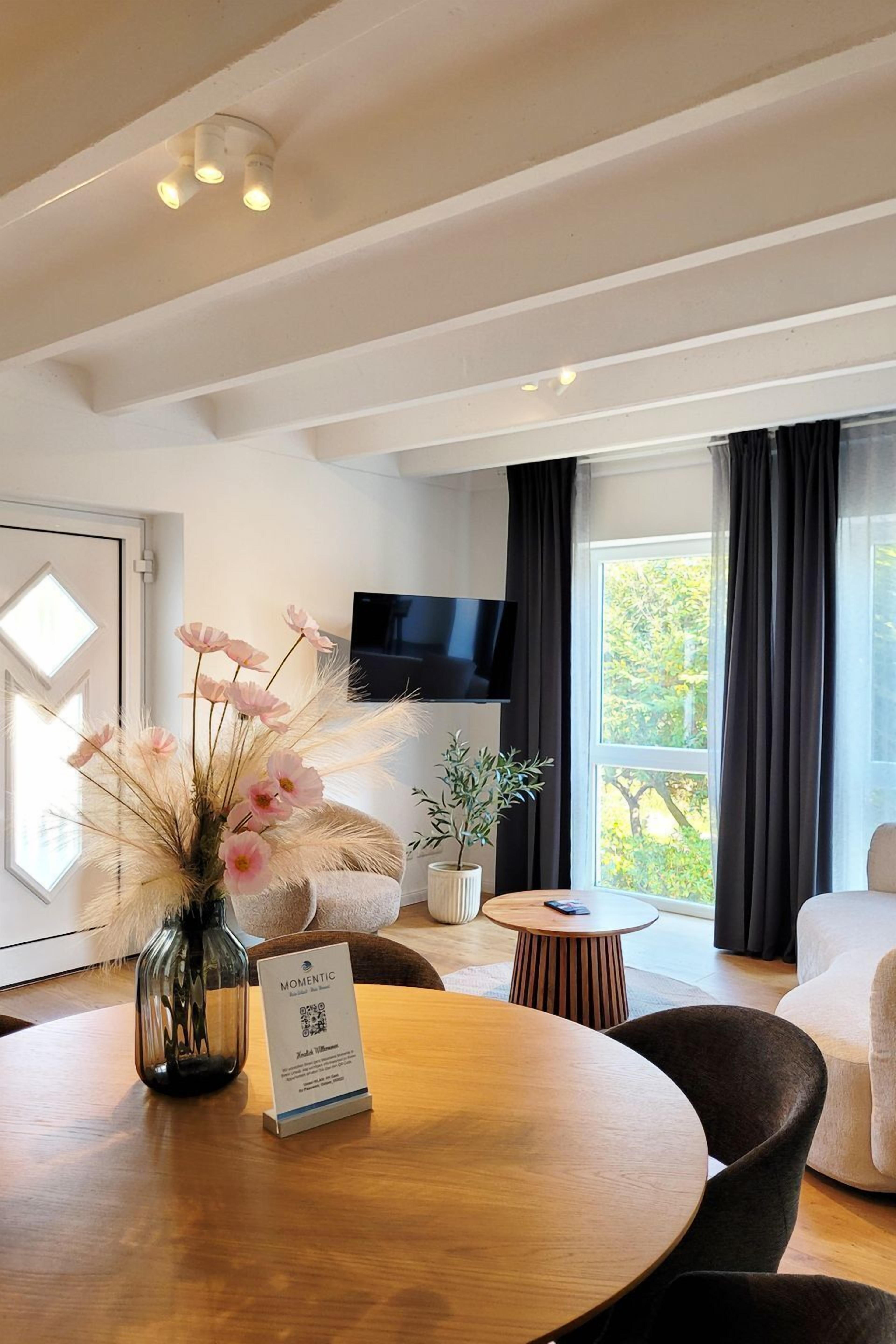 Une chambre d’hôtel moderne avec une table ronde en bois, un vase rempli de fleurs roses, une petite plante en pot, un téléviseur à écran plat fixé au mur et une grande fenêtre avec des rideaux.