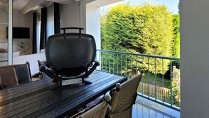 A balcony with a grill, a dining table, chairs, and a view of a green hedge.