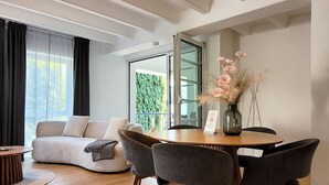 A modern living room with a sofa, a dining table with chairs, a vase with flowers, and a large window with a view of greenery.