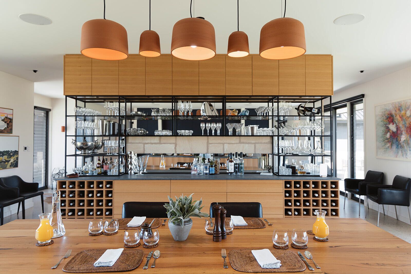 A modern dining area with a wooden table set for a meal, a bar area with shelves of glasses and bottles, and pendant lighting.