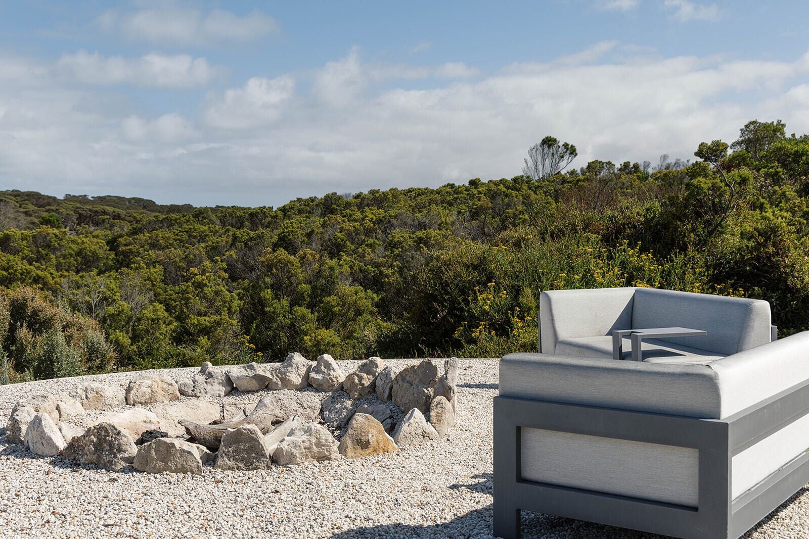 Un espacio exterior moderno con asientos, una chimenea de piedra y un sofá, frente a un fondo de vegetación densa y colinas.