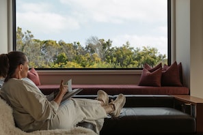 A person sitting on a sofa with a laptop, a window with a view of trees, and a red pillow on the sofa.