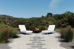 A patio with two white lounge chairs and a central fire pit, surrounded by a gravel path and greenery.