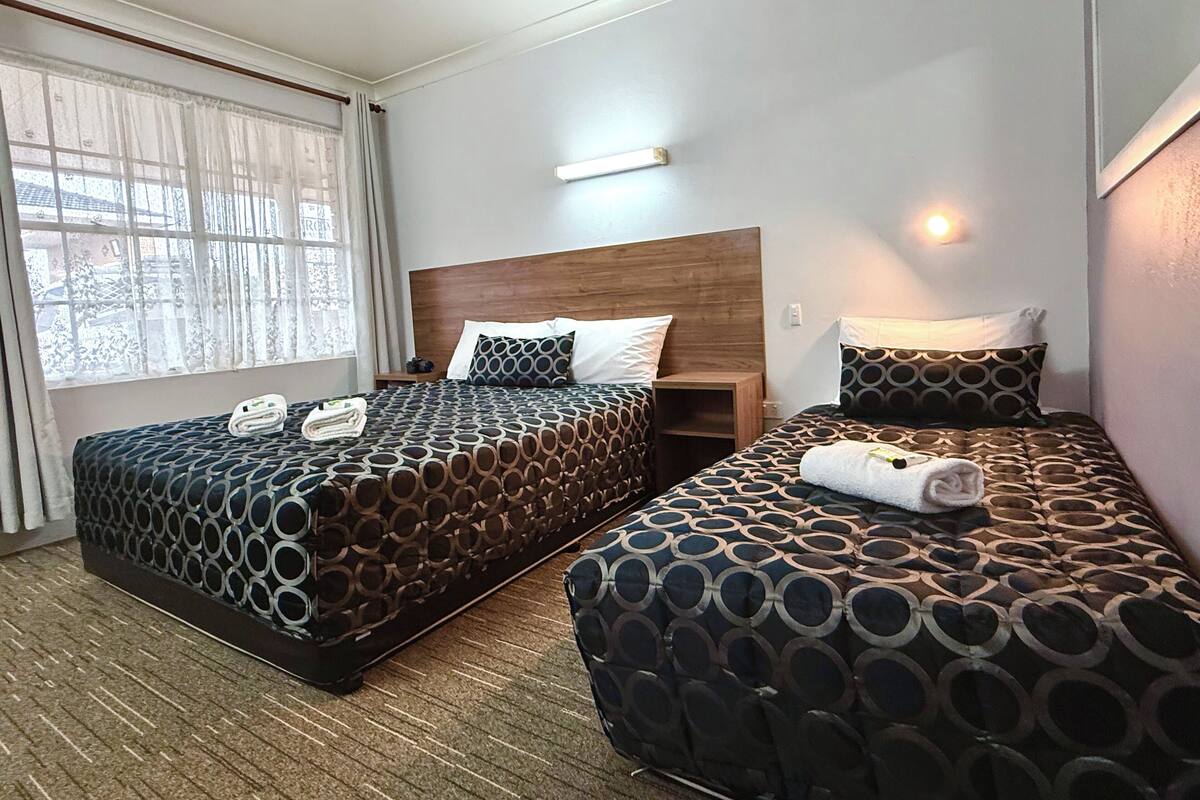 Habitación de hotel con dos camas, cada una con colcha y almohadas estampadas, cabecero de madera, mesita de noche y ventana con cortinas traslúcidas.