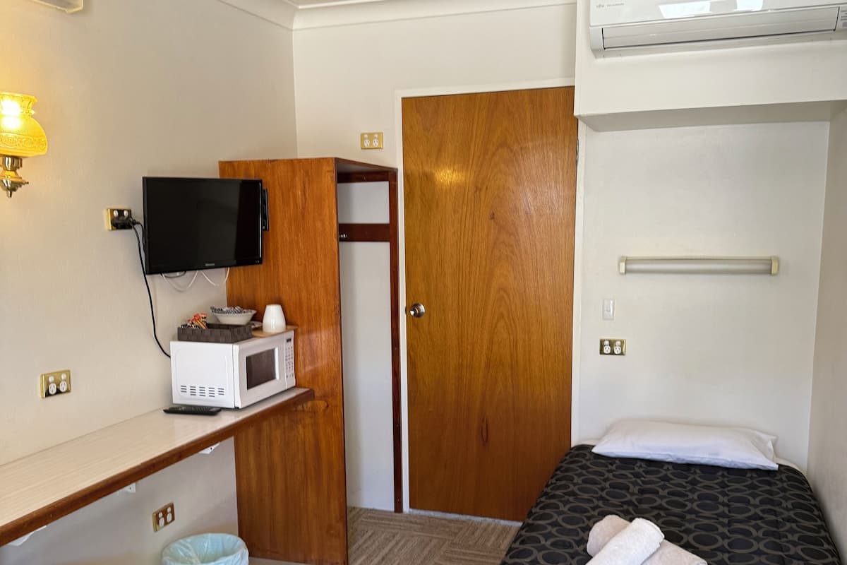 Habitación de hotel con cama, escritorio con televisor, microondas, armario de madera y cubo de basura.