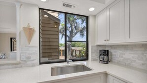 A kitchen with a large window, white cabinets, a stainless steel sink, and a coffee maker.