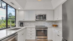 A modern kitchen with white cabinets, a stainless steel oven, a microwave, and a large window offering a view of greenery.