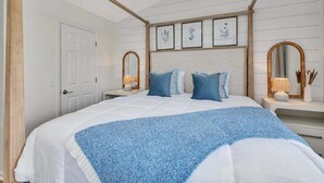 A bedroom with a large bed, two bedside tables with lamps, a wooden headboard, and framed botanical prints on the wall.
