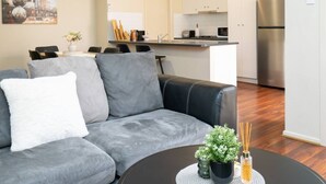 A modern living room with a grey sofa, a small round table with a plant, and a kitchen in the background.