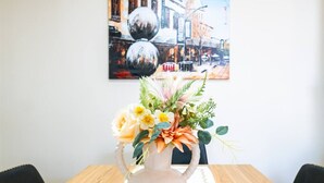 A dining table with a white vase of flowers, a painting of a street scene, and a wooden table.