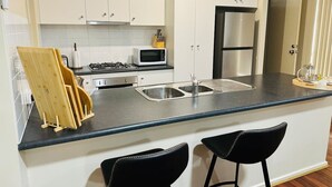 A modern kitchen with white cabinets, a stainless steel sink, a microwave, a gas stove, and two black bar stools.