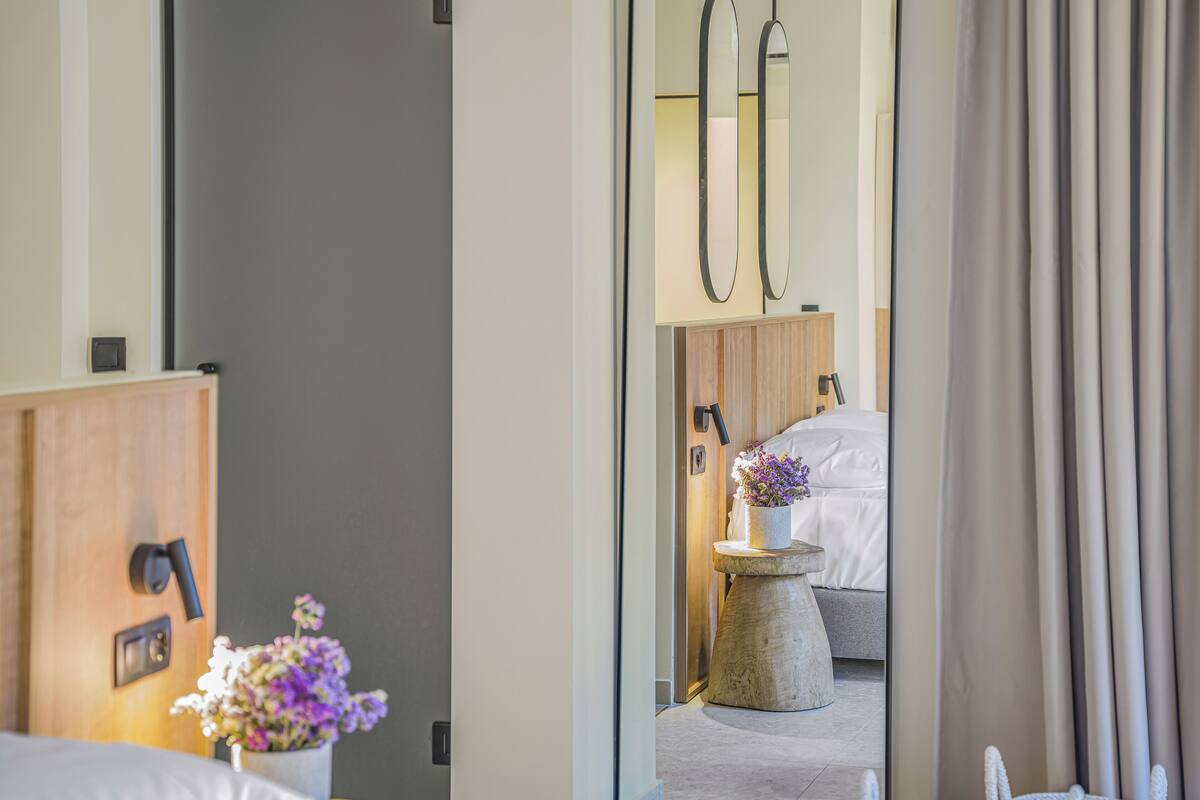 A hotel room with a wooden wardrobe, a bedside table with a vase of flowers, a bed with a grey headboard, and a small round table with a lamp.