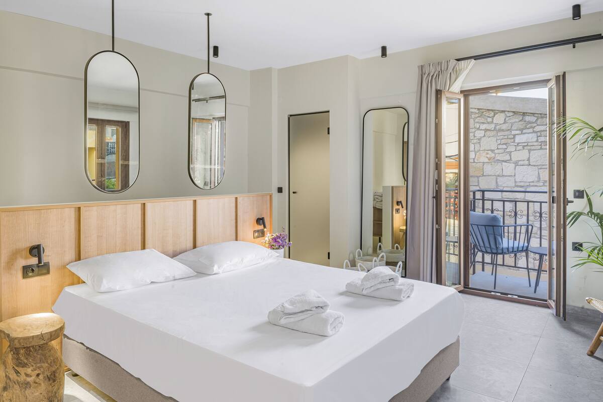A modern hotel room with a large bed, two bedside tables, and a balcony with a view of the stone wall and outdoor seating.