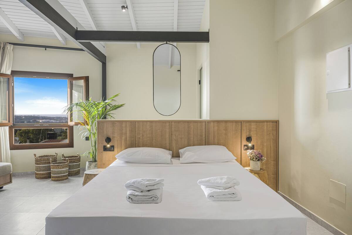 A modern hotel room with a large bed, a wooden headboard, a large window with a view, and a potted plant.