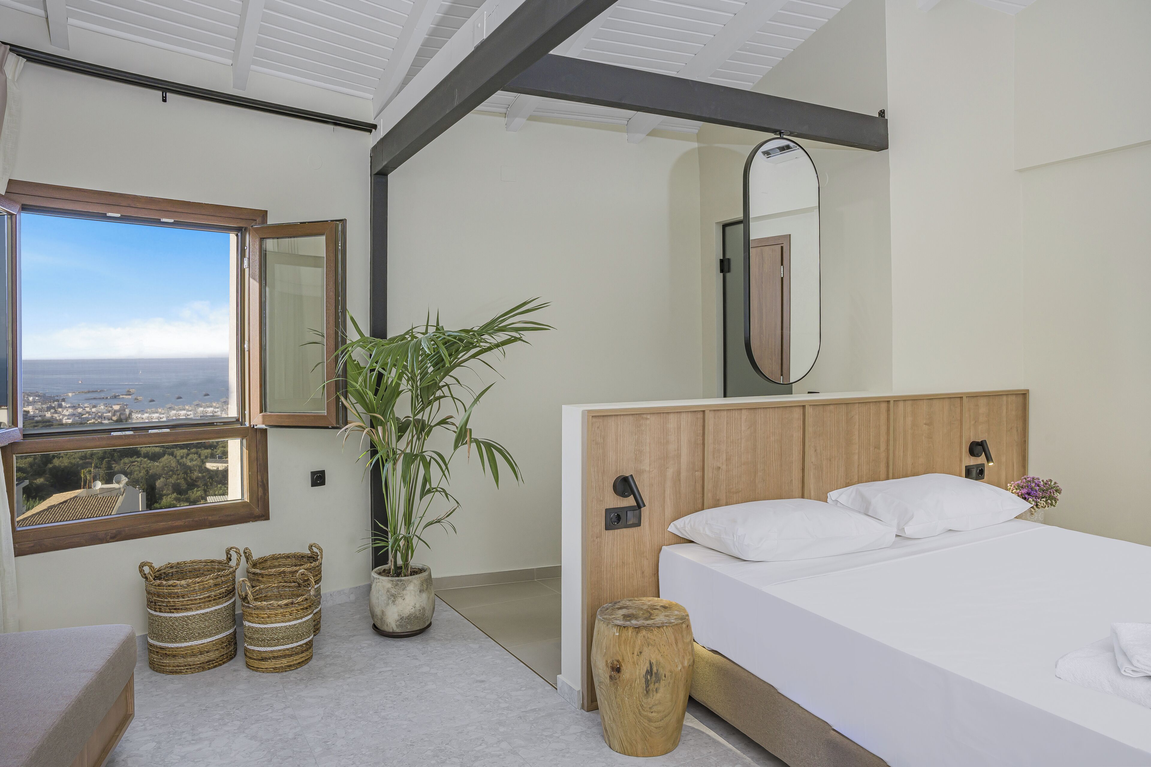 A bedroom with a large window, a bed, a bedside table, a mirror, and a potted plant.