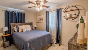 A bedroom with a bed, a ceiling fan, a nightstand, a lamp, a rug, a wooden dresser, and a wall-mounted shelf with decorative items.