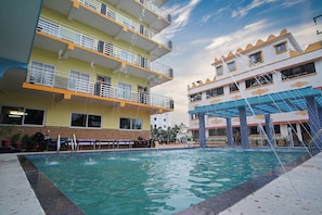 A swimming pool with water fountains, surrounded by multi-story buildings with balconies and outdoor seating areas. - HOTEL DREAM BASANTI (Kanthi)