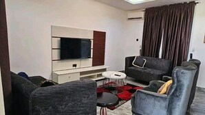 A modern living room with a flat-screen TV mounted on a white wall, a grey sofa, a small round coffee table, and a grey armchair. - Geoworld Apartments & Suites (Makurdi)