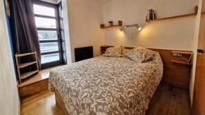 A bedroom with a large bed, a window with curtains, a bedside table, a nightstand, and a wall-mounted shelf.