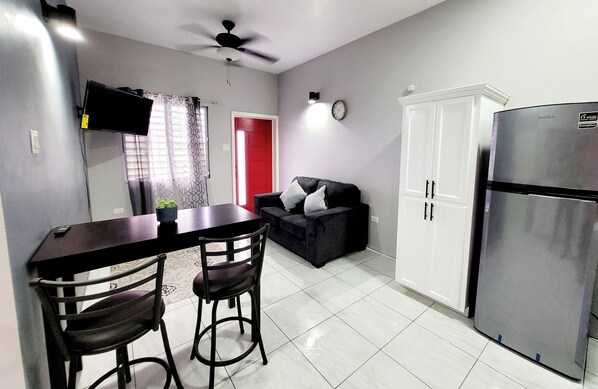 A compact kitchen with a dining area, a refrigerator, a ceiling fan, a TV mounted on the wall, and a red door.