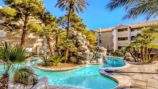 A swimming pool with a natural rock feature, surrounded by palm trees and a building with a tiled roof. - Upgraded Vegas Oasis! Heated Pool, Hot Tub & Gym! (Las Vegas)