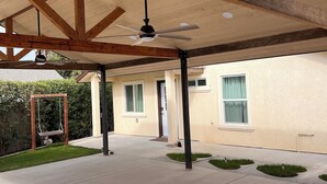 Un espacio al aire libre techado con suelo de concreto, vigas de madera, un ventilador de techo y un conjunto de columpios.