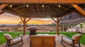Un patio exterior con zona de estar techada, una parrilla y vistas a un paisaje montañoso al atardecer.
