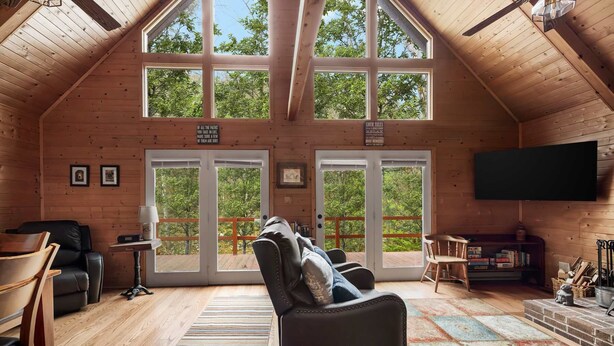 A cozy cabin with a high ceiling, wooden walls, and large windows offering a view of the trees. There is a comfortable seating area with a sofa and chairs, a flat-screen TV mounted on the wall, and a bookshelf with books and decorative items. - Loft, Wood Stove, Fire Pit, Grill & Deck on Creek (Hedgesville)