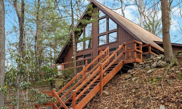 A cabin with a wooden staircase leading to a large window, surrounded by trees. - Loft, Wood Stove, Fire Pit, Grill & Deck on Creek (Hedgesville)