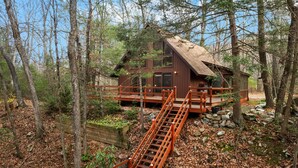 A cabin with a wooden deck and a ladder leading to a wooden staircase. - Loft, Wood Stove, Fire Pit, Grill & Deck on Creek (Hedgesville)