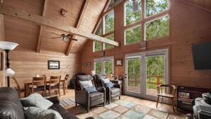 A cozy cabin interior with wooden walls, a ceiling fan, a fireplace, and a large window offering a view of the outdoors. - Loft, Wood Stove, Fire Pit, Grill & Deck on Creek (Hedgesville)