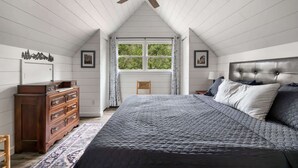 A bedroom with a large bed, wooden dresser, ceiling fan, and a window with curtains. - Loft, Wood Stove, Fire Pit, Grill & Deck on Creek (Hedgesville)