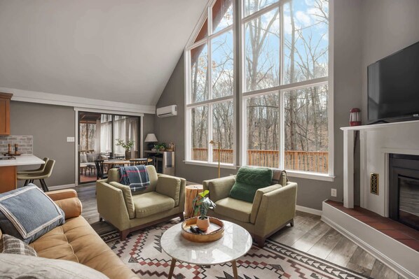 A spacious living room with a high ceiling, large windows, a fireplace, and a dining area visible through an open door.