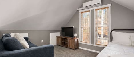 A bedroom with a sloped ceiling, a TV on a wooden stand, a bed with white bedding, and a window with blinds.