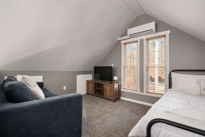 Un dormitorio con techo inclinado, un televisor sobre un soporte de madera, una cama con ropa blanca y una ventana con persianas.