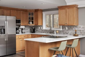 A modern kitchen with wooden cabinets, a stainless steel refrigerator, a large island with bar stools, and a window with a view of trees.