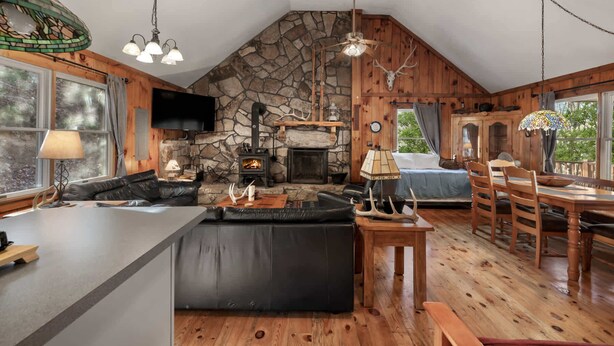 A rustic cabin interior with a stone fireplace, wooden floors, a large island in the center, and a dining area with wooden furniture. - Rustic retreat w/ kitchen & deck (Great Cacapon)
