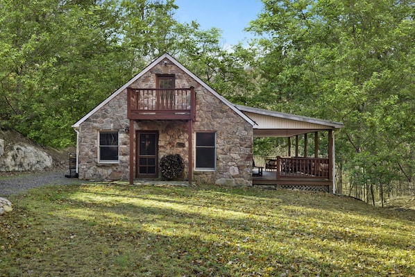 A stone cabin with a balcony, a covered porch, and a grassy yard surrounded by trees.